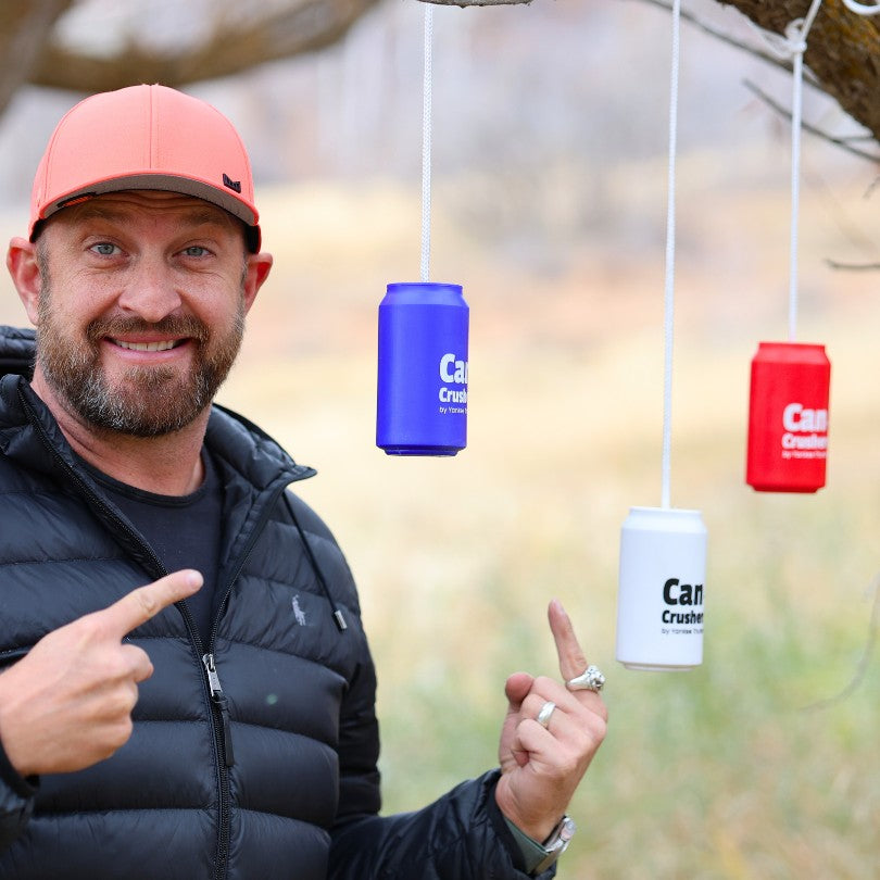 A man in a red cap and black jacket stands outdoors, smiling and pointing at three hanging Yankee Thunder Can Crushers Targets ⭐️⭐️⭐️⭐️⭐️ in blue, white, and red. The targets are suspended from a tree branch. The background features a blurred, natural setting.