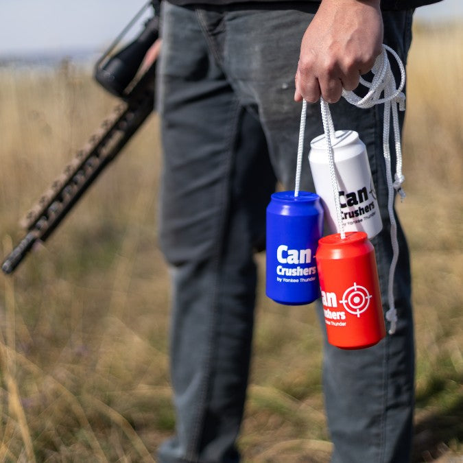 A person is holding a rifle in one hand and four colorful, shatterproof Yankee Thunder Can Crushers Targets ⭐️⭐️⭐️⭐️⭐️ in the other. Standing in a grassy outdoor setting, the durable and reusable cans are connected by a white rope.
