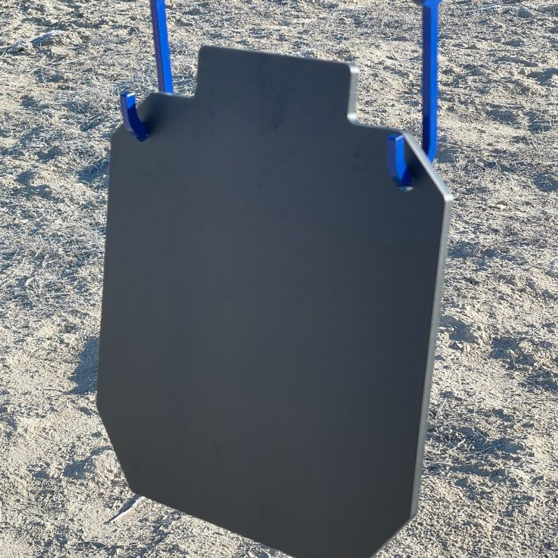 A close-up of a Yankee Thunder 14 Inch Silhouette Body Mass Target hanging from a blue metal frame, set up outdoors on a sandy surface. The unmarked rectangular target has a small notch at the top, making it ideal for shooting practice and as a training aid.