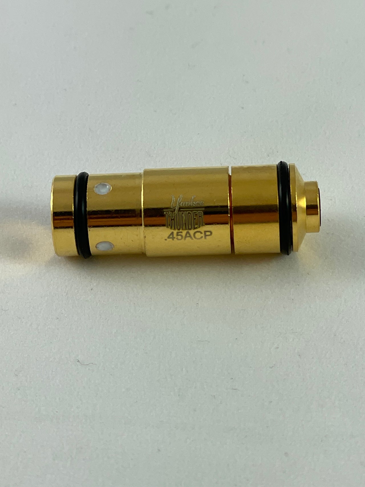 A close-up photograph of a golden .45 Laser Cartridge by Yankee Thunder with the text ".45 ACP" and "Laser Thunder" etched on it. The casing, ideal for dry-fire practice, features two black O-rings near each end and is lying on a plain, light-colored surface.