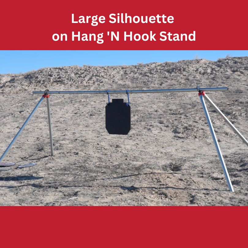 A large 14 Inch Silhouette Body Mass Target from Yankee Thunder hangs from a metal Hang 'N Hook stand set up on a dirt and rock terrain. The stand has three legs, two on the left and one on the right, providing stability. The background features a rocky, arid landscape under a clear sky, perfect for shooting practice.