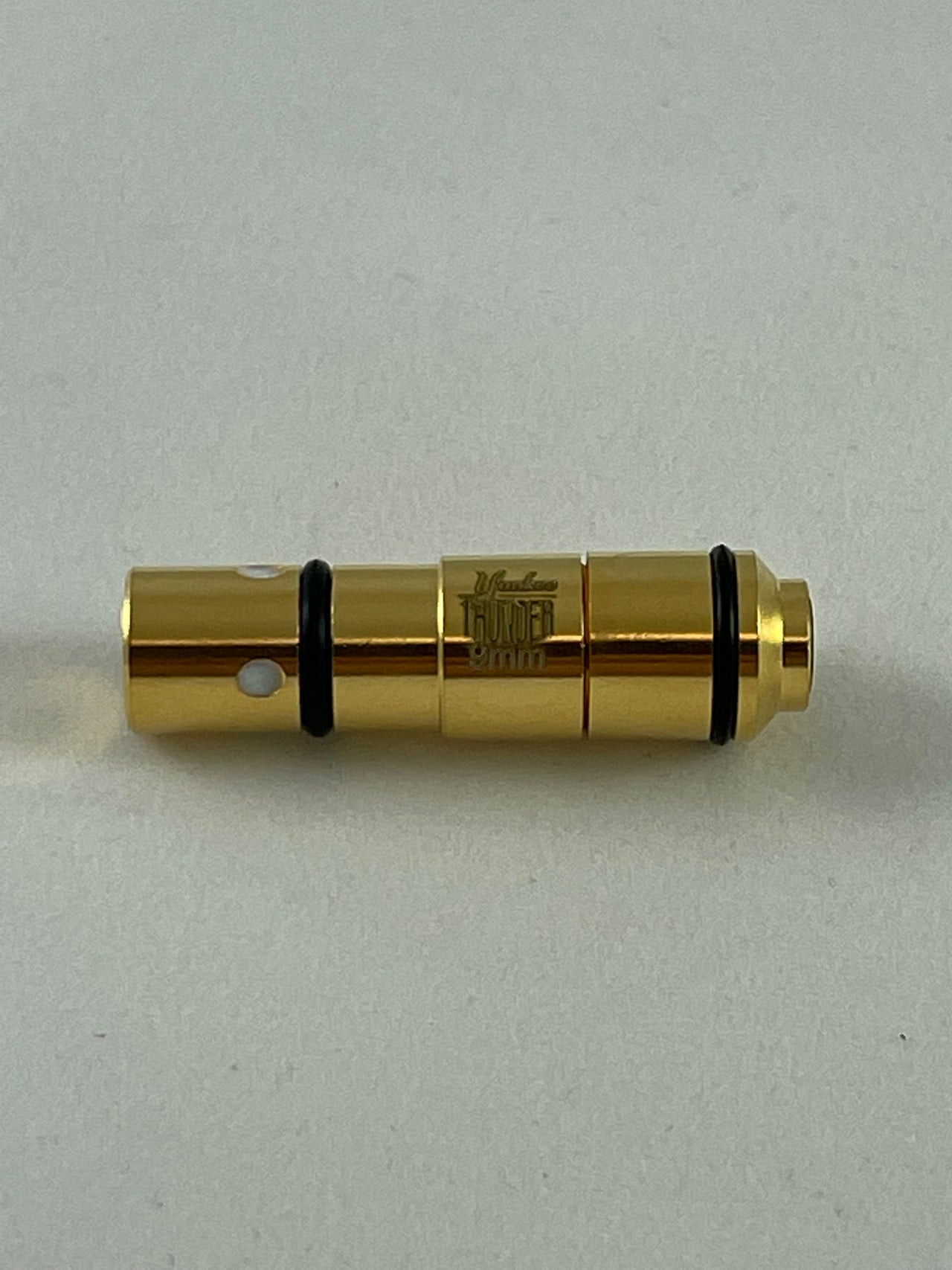 A close-up view of the Yankee Thunder 9mm Laser Cartridge, a golden cylindrical object with black rings, resembling a precision-engineered part or connector. This item, which is designed for dry-fire practice, features engraved text and has visible holes near one end. It is lying on a plain, light-colored surface.