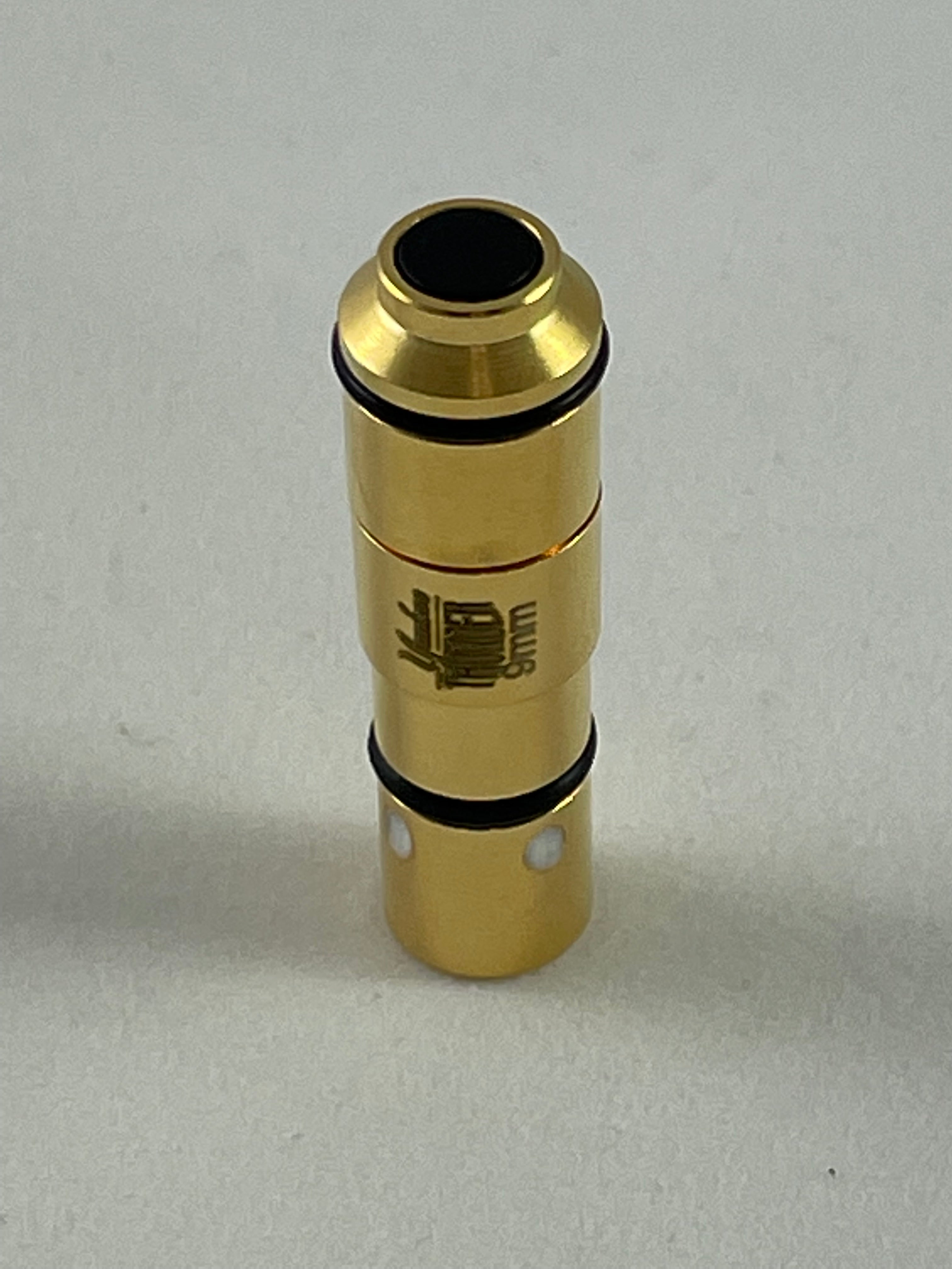 A Yankee Thunder 9mm Laser Cartridge, gold-colored with black ring accents and resembling a small mechanical part or connector, stands vertically on a neutral surface. It has a small black circular opening at the top and features an engraved emblem or design on its side. Ideal for laser cartridge dry-fire practice with the Straight Shooter Laser Training App.