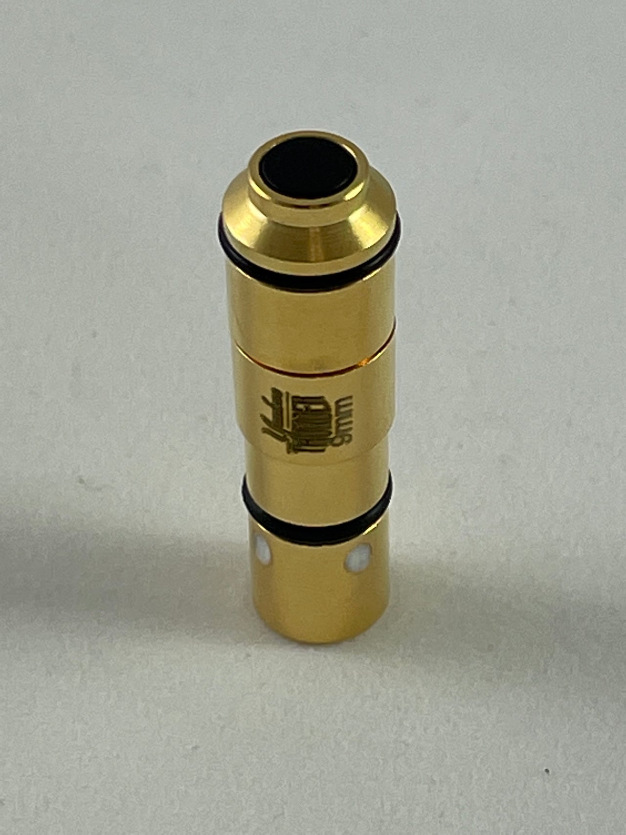 A Yankee Thunder 9mm Laser Cartridge, gold-colored with black ring accents and resembling a small mechanical part or connector, stands vertically on a neutral surface. It has a small black circular opening at the top and features an engraved emblem or design on its side. Ideal for laser cartridge dry-fire practice with the Straight Shooter Laser Training App.