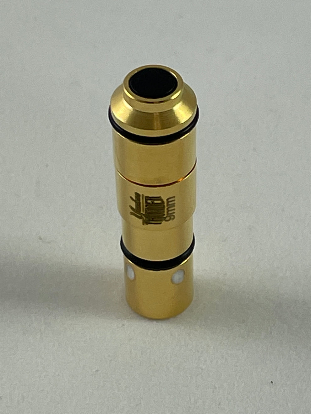 A Yankee Thunder 9mm Laser Cartridge, gold-colored with black ring accents and resembling a small mechanical part or connector, stands vertically on a neutral surface. It has a small black circular opening at the top and features an engraved emblem or design on its side. Ideal for laser cartridge dry-fire practice with the Straight Shooter Laser Training App.