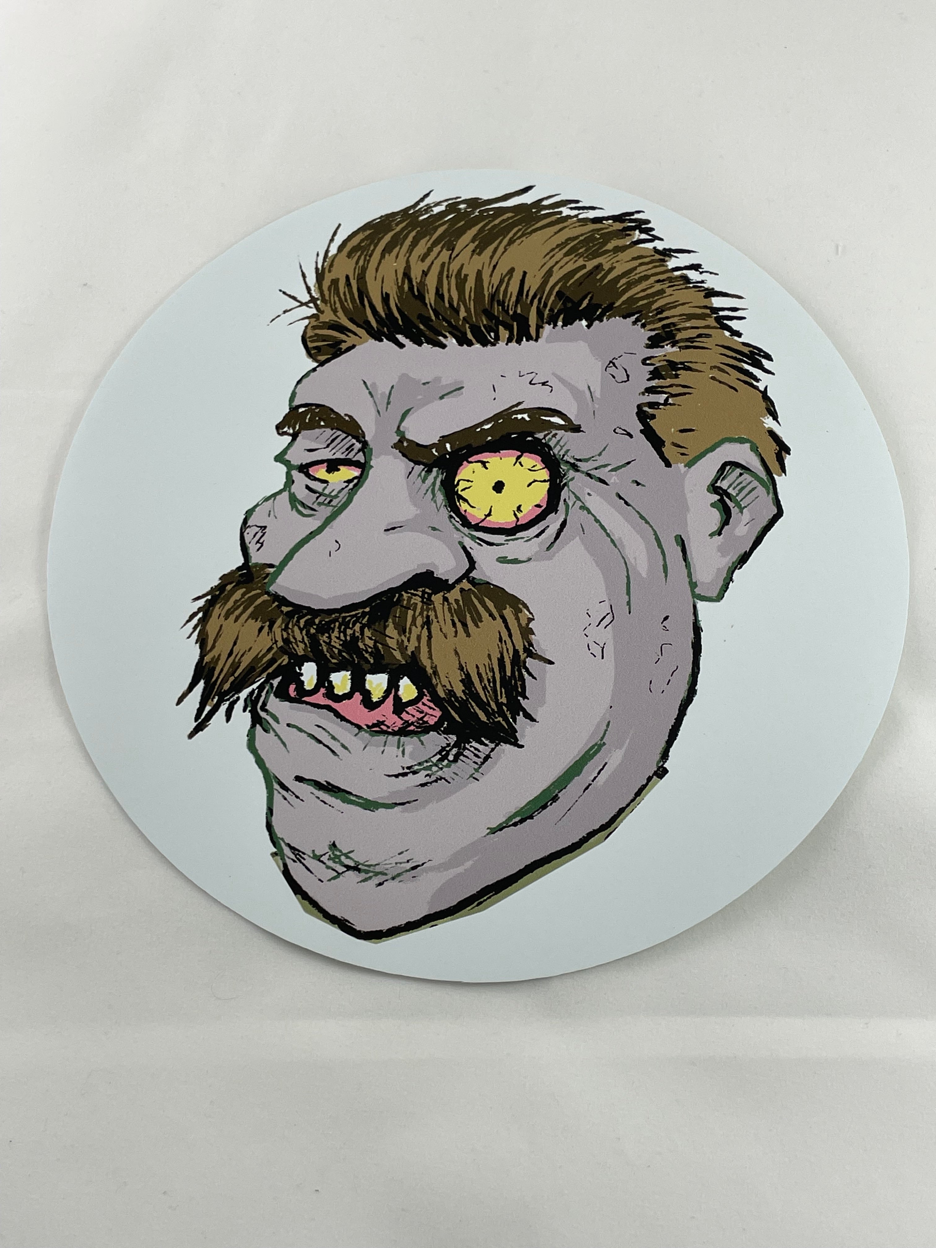 Illustration on an 8-inch Zombie Target Sticker by Yankee Thunder, featuring a menacing, zombie-like face with yellow, bloodshot eyes, uneven, sharp teeth, and an exaggerated mustache. The face has a grayish complexion and wild, disheveled hair set against a pale background—perfect for stickers featuring notorious villains in history.